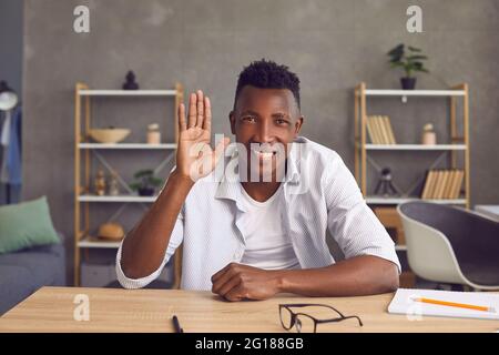Happy black student sitting at his desk at home, smiling and waving hand at camera Stock Photo