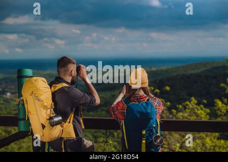 Couple of young hikers taking photo with a smartphone and looking into nature with a binoculars while standing on a lookout Stock Photo