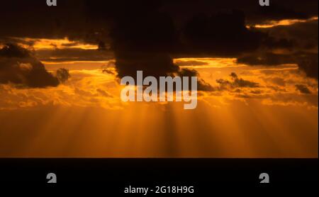 Sunset over the ocean with god rays shining through dark threatening clouds and orange sky. Stock Photo