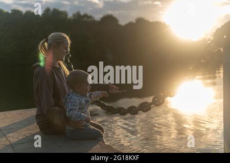 Mom and son with blond hair sit on the bridge to the shore of the lake during sunset. Stock Photo