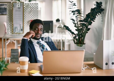 Stylish black male worker talking on cellphone at table with netbook and hot drink to go while looking at camera Stock Photo