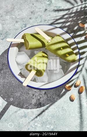 Frozen homemade pistachio popsicle in bowl of ice on gray background with palm leaf shadow in harsh light. Refreshing popsicle, frozen green juice on Stock Photo