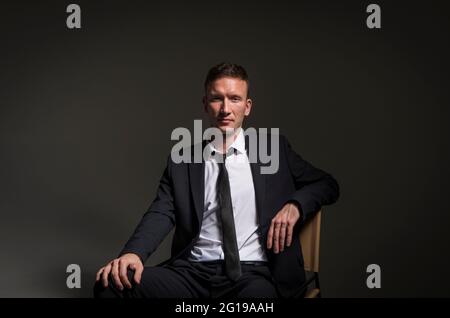 Portrait of a pensive confident man, in a business suit, sitting cross-legged against a dark background. Stock Photo
