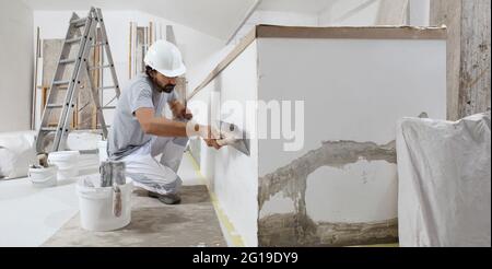 man plasterer construction worker at work, takes plaster from bucket and puts it on trowel to plastering the wall, wears helmet inside the building si Stock Photo