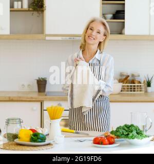 Charming senior woman in apron enjoying cooking on retirement in light modern kitchen at home, wiping hands with towel. Smiling retired 60s female looking at camera with smile while preparing food Stock Photo