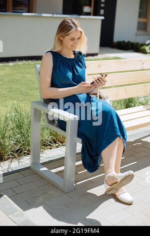 Young beautiful girl sits on bench in city, smiles and uses smartphone. Concept of new technologies and communication on the Internet Stock Photo