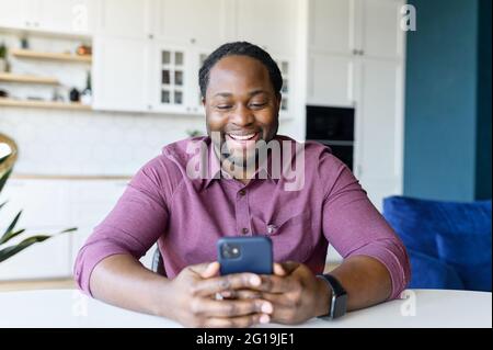 Positive African-American guy enjoys spending time online in social networks, sitting at the desk at home and using smartphone, messaging and chatting, black man holds mobile and laughs Stock Photo