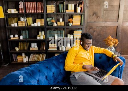 African American male student freelancer wear yellow hoodie learn on-line use wireless headphones, laptop sit at sofa at modern home. Young Stock Photo