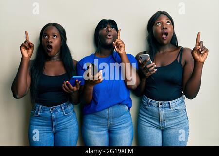 Three young african american friends using smartphone amazed and surprised looking up and pointing with fingers and raised arms. Stock Photo
