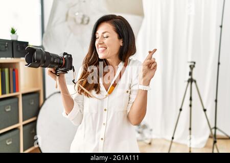 Beautiful caucasian woman working as photographer at photography studio gesturing finger crossed smiling with hope and eyes closed. luck and superstit Stock Photo