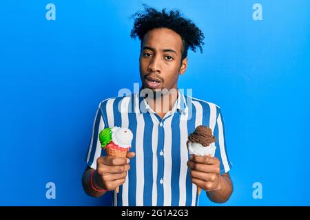 Young african american man with beard holding two ice cream cones in shock face, looking skeptical and sarcastic, surprised with open mouth Stock Photo