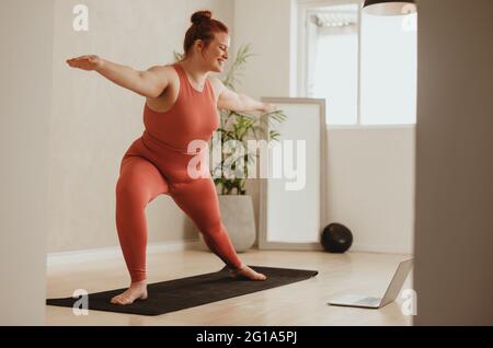 Smiling woman exercising at home and watching training videos on laptop. Plus size female doing yoga with hands outstretched and looking at laptop. Stock Photo