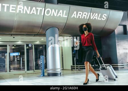 Businesswoman wearing face mask with luggage walking in airport terminal. International traveler arrived with suitcase during pandemic. Stock Photo