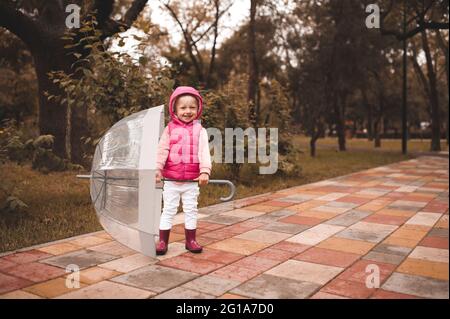 Cute child girl 3-4 year old holding umbrella wear vest with hood and rain rubber boots in park outdoors. Little toddler having fun outside. Cheerful Stock Photo