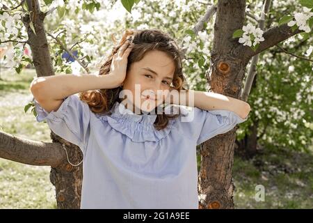 Lifestyle photo of a happy child. Cute pretty smiling brunette girl in a blooming garden. The girl straightens her fluttering hair in the wind and loo Stock Photo