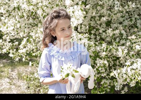 Lifestyle photo happy child. Cute pretty smiling brunette girl with a favorite soft toy in her hands. A girl stands against a background of blooming a Stock Photo