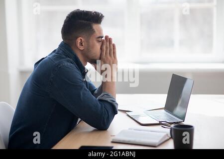 Exhausted arab man sitting in front of laptop, office interior Stock Photo