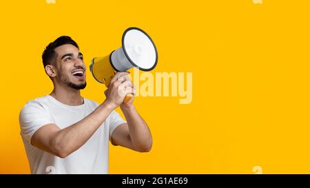 Excited arab guy speaking loud, using megaphone on yellow Stock Photo