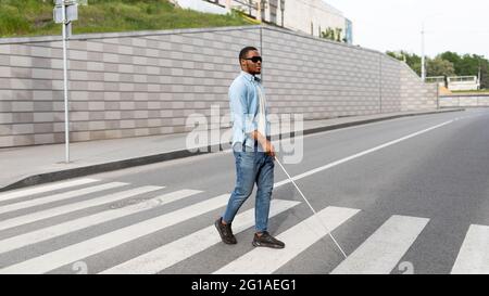 Young black visually impaired man wearing dark glasses, walking across city street, using cane Stock Photo