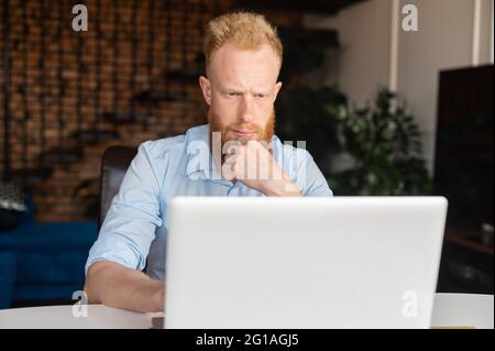 Absorbed and thoughtful red haired businessman staring at the computer monitor, concentrated ginger head freelancer using laptop in the home office, solving tasks, browse internet, researching topic Stock Photo