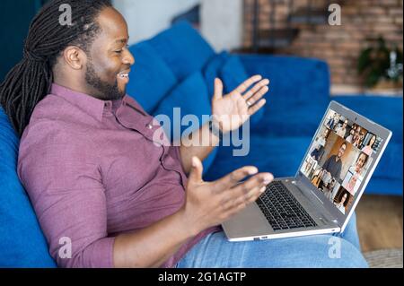 Happy African-American guy makes video call on the laptop, talking and discussing with group of diverse colleagues on the distance, using new computer app for video connection, takes part in webinar Stock Photo