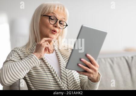 Surprised mature woman using tablet sitting on couch Stock Photo
