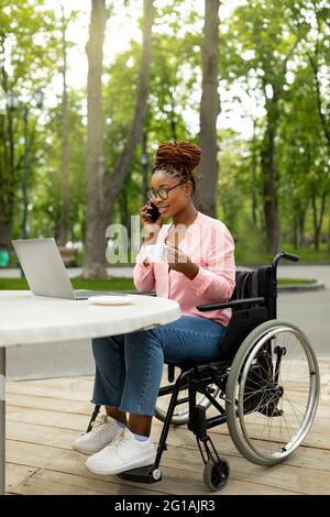 Happy black lady in wheelchair using laptop, speaking on smartphone at outdoor cafe Stock Photo