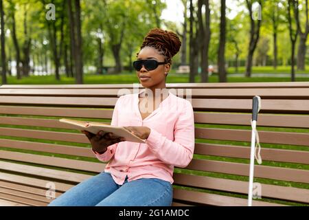 Millennial black visually impaired woman sitting on bench at city park, reading Braille book outside Stock Photo