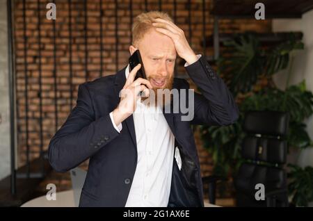 Disappointment redhead businessman talking on the smartphone, ceo has phone conversation in modern office, received bad news, bankruptcy, money losing, man in formal suit holding head, feels despair Stock Photo