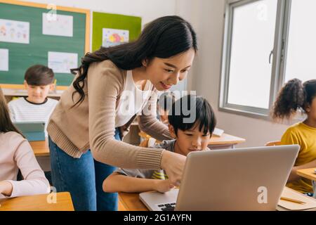 Young adult smiling beautiful Asian teacher helping elementary student boy with laptop in computer classroom. Information Technology class in primary school concept. Stock Photo