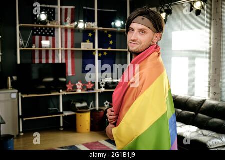Young gay man with pride flag standing in living-room and looking at camera Stock Photo