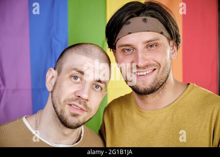 Two happy gay men standing against rainbow flag and looking at you Stock Photo