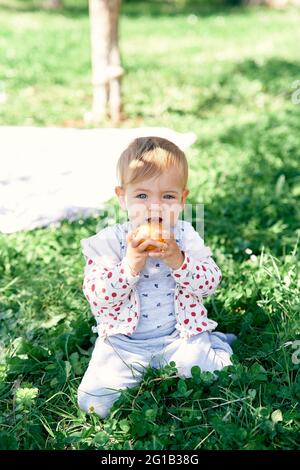 Cute baby sitting on green grass and gnawing a yellow apple Stock Photo