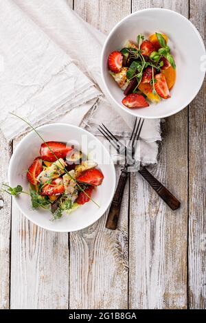 Delicious summer salad. Salad with fresh strawberries, baked beets, yellow tomatoes, dor blue cheese and herbs in a white bowl on a light rustic backg Stock Photo