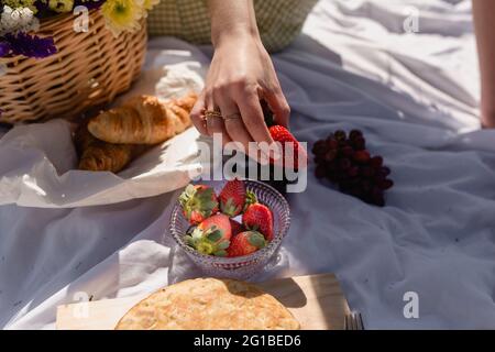 High angle of crop female with ripe strawberry having picnic with croissant and grapes with focaccia Stock Photo