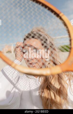 Positive young female in white clothes touching hair and looking at camera through tennis racket while standing on sunny street Stock Photo