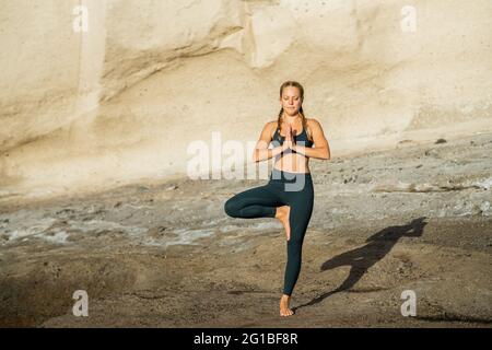 Young mindful female in sportswear on vrksasana looking down while practicing yoga standing on rocky background Stock Photo