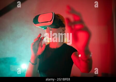 Happy Woman wearing t shirt while using VR goggles and standing in studio with red neon lights Stock Photo