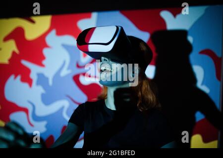 Happy lady wearing black t shirt while standing in room with colorful lights and using VR headset Stock Photo