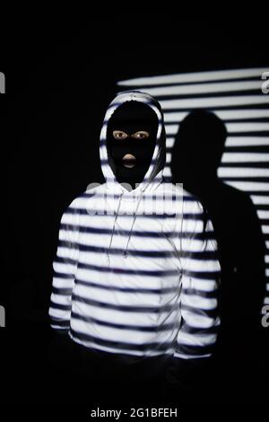 Unrecognizable serious rebellious young male in black balaclava mask and white hoodie standing near wall with jalousie shadow Stock Photo