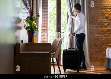 Full body low angle side view of adult man with luggage opening curtain hanging on balcony door in hotel room Stock Photo