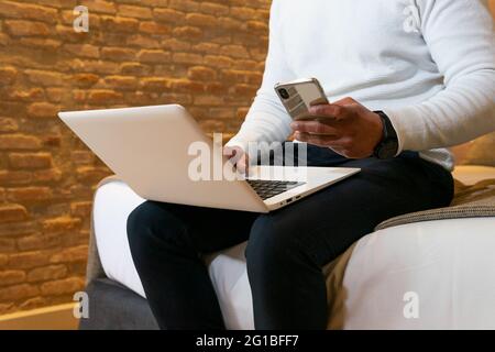 Low angle of crop ethnic male entrepreneur sitting on bed and browsing mobile phone and working on laptop Stock Photo