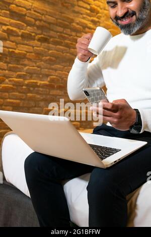Low angle of crop bearded ethnic male entrepreneur sitting on bed and having drink while browsing mobile phone and working on laptop Stock Photo