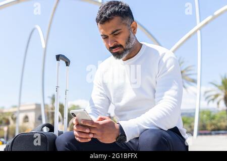 Concentrated adult male in smart casual clothes browsing mobile phone and sitting on sunny street with suitcase Stock Photo
