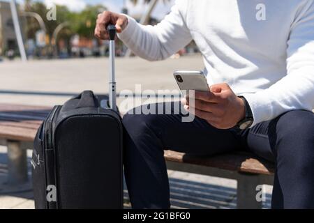 Crop male in smart casual clothes browsing mobile phone and sitting on sunny street with suitcase Stock Photo