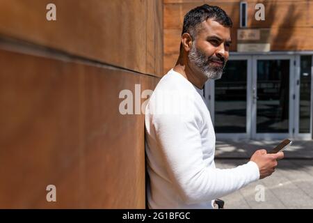 Side view glad bearded male in white sweater standing near modern building wall and looking away in thoughts on sunny day Stock Photo