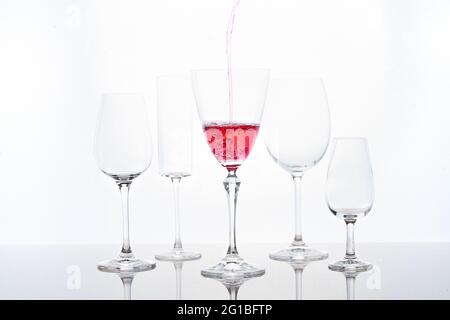 Anonymous person pouring red sparkling wine into goblet laced on mirrored table near various shaped glasses against white background Stock Photo