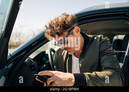 Serious male in sunglasses browsing cellphone while sitting in automobile on sunny day Stock Photo