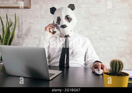 Unrecognizable male entrepreneur in panda bear mask speaking on cellphone against netbook during telework at desk Stock Photo