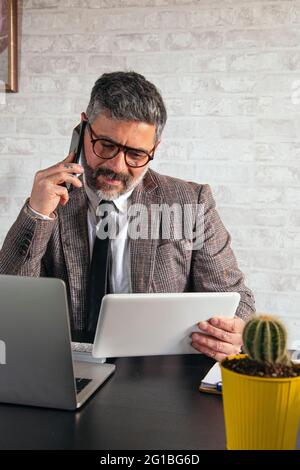 Mature male entrepreneur with tablet talking on cellphone at table with netbook in loft style office Stock Photo